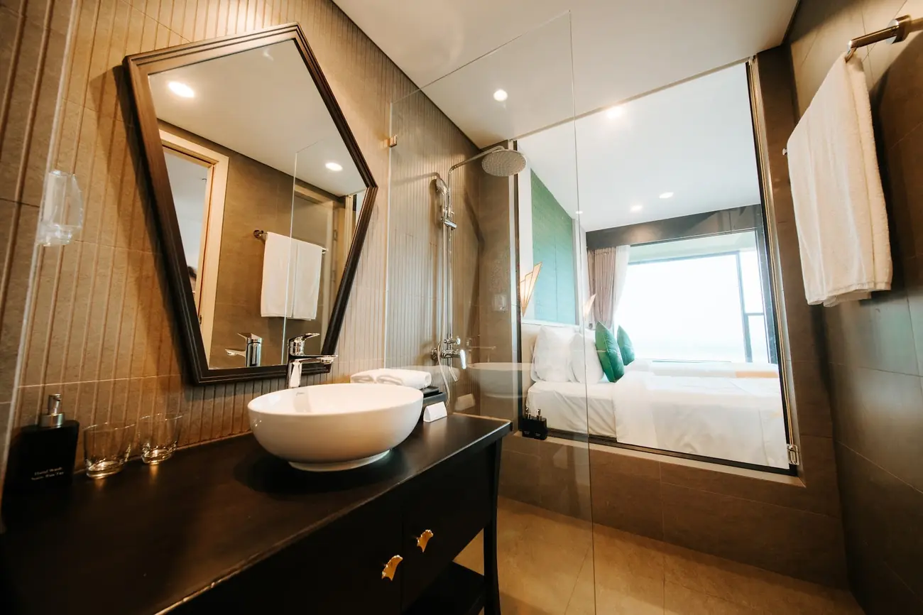 Phòng tắm phòng Deluxe với gương lục giác và vách kính nhìn vào phòng ngủ – khách sạn Lynntimes Thanh Thủy